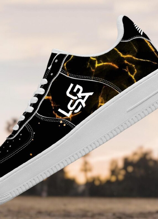 The LIONSGALA - LSGA Dorado Bolted Gold streetwear sneakers by LIONSGALA Brands feature a black upper, white soles and laces, a yellow lightning pattern, and bold white side logo, shown against a blurred outdoor backdrop.