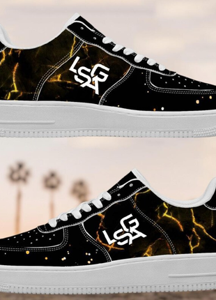 A pair of LIONSGALA - LSGA Dorado Bolted Gold streetwear sneakers by LIONSGALA Brands, featuring a black upper with a gold marble pattern, white soles, and white LSR logos. The background is blurry with palm trees and a soft sunset sky.