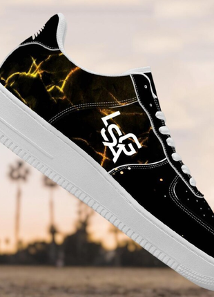 The LIONSGALA - LSGA Dorado Bolted Gold streetwear sneakers by LIONSGALA Brands feature a black design with white laces and sole, gold lightning accents, and bold “LSR” lettering on the side, set against palm trees in the background.