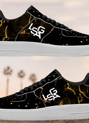 Two black LIONSGALA LSGA Dorado Bolted Gold streetwear sneakers by LIONSGALA Brands, with white soles and laces, gold lightning pattern, and white LSR logo on the side, set against a blurred background with faint palm trees.