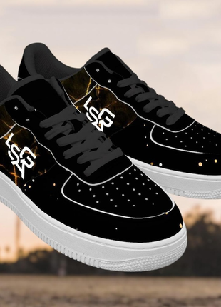 The LIONSGALA - LSGA Dorado Bolted Gold streetwear sneakers by LIONSGALA Brands feature a black design with white soles and laces, bold LSR branding, and a striking gold marble-like pattern, shown outdoors against a blurred background.
